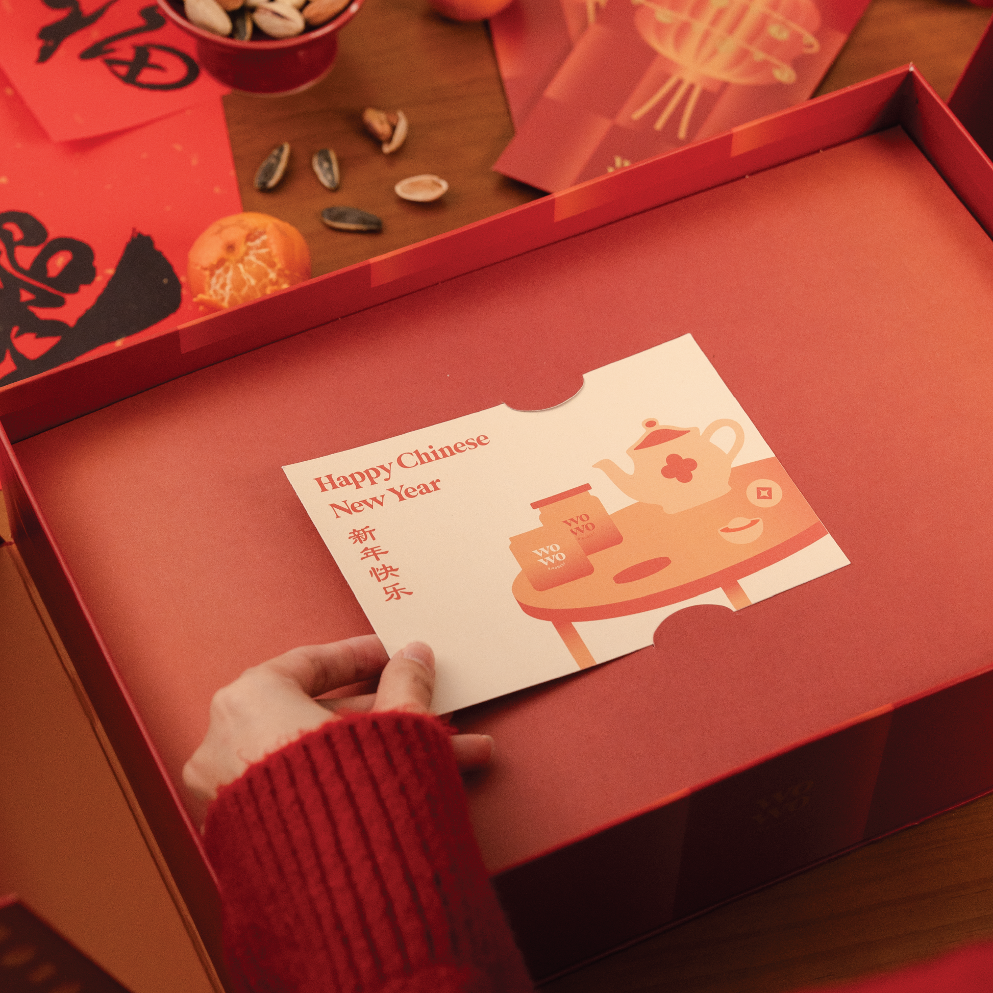 Person holding a 'Happy Chinese New Year' card over a red box with festive decorations.